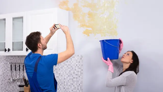 Homeowner holding a bucket under a ceiling leak while a technician photographs the damage
